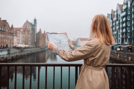 Téléchargez les photos : Attrayant jeune touriste féminine explore la nouvelle ville. Fille blonde tenant une carte sur papier sur la rue de la ville à Gdansk. Voyager en Europe en automne. - en image libre de droit