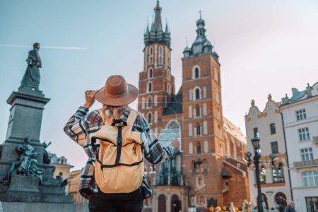 Téléchargez les photos : Vue arrière de la femme voyageuse marchant sur la vieille place du marché à Cracovie tenant carte touristique. Concept de voyage et mode de vie actif. Photo de haute qualité - en image libre de droit