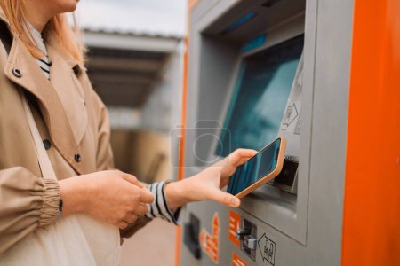 A side view of a young beautiful elegant blonde woman paying for service underground parking or buying a subway or train ticket using an electronic self-service kiosk. Travel, airport and couple at
