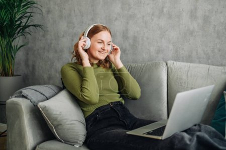 Headphones, laptop and woman on a home sofa listening to music or audio while streaming online. Calm female person relax on couch to listen to radio or song with internet connection and technology