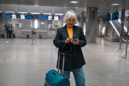 Photo for Smiling 60s senior business woman passenger in black jacket with suitcase using phone in terminal hall while waiting for her flight. Application for travel, banking, concept. - Royalty Free Image