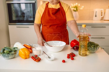 Téléchargez les photos : Femme assaisonne une salade de légumes tout en préparant la nourriture dans la cuisine. Chef méconnaissable ajoute de l'huile d'olive tout en préparant un repas végétarien à la maison. Concept de saine alimentation. - en image libre de droit