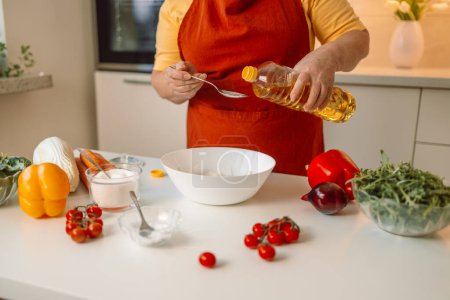 Téléchargez les photos : Femme assaisonne une salade de légumes tout en préparant la nourriture dans la cuisine. Chef méconnaissable ajoute de l'huile d'olive tout en préparant un repas végétarien à la maison. Concept de saine alimentation. - en image libre de droit