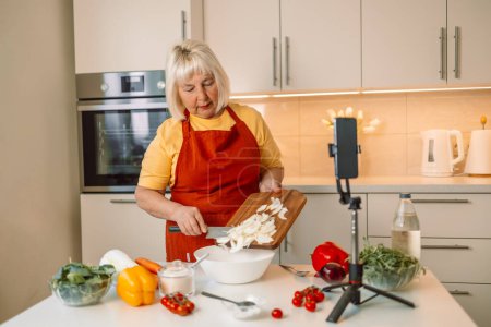 Téléchargez les photos : Blogueuse alimentaire caucasienne travaillant sur une nouvelle vidéo et expliquant comment cuisiner un plat. Mains de femmes âgées des années 60 hachant l'oignon frais sur une planche de bois tout en préparant des pâtes italiennes aux légumes - en image libre de droit