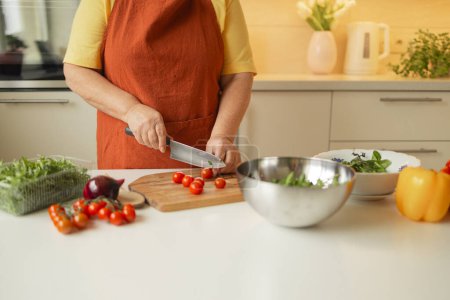 Female chef is precisely slicing fresh cherry tomato on wooden board while preparing Italian salad with vegetables in kitchen