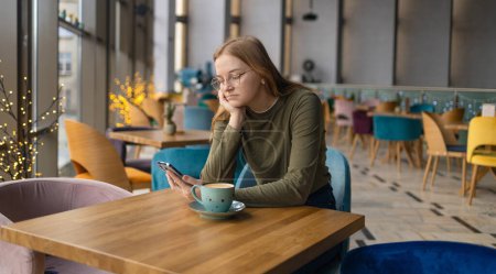 Mujer joven triste y pensativa que bebe café y mira su teléfono inteligente cerca de la ventana de la cafetería. Foto de alta calidad