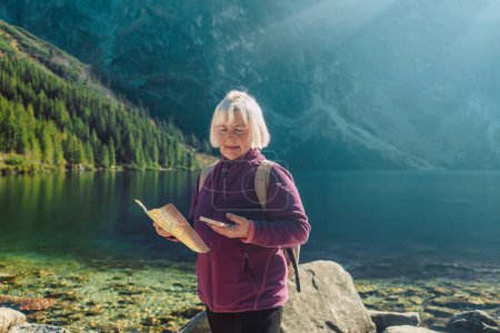 Aufnahme einer Seniorin um die 60, die mit ihrem Smartphone eine Landkarte hält, während sie die Aussicht von der Spitze eines Berges genießt. Touristinnen sitzen auf einem Felsen in den Bergen und lesen die Landkarte. Polen, Tatry 