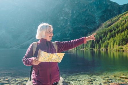 Shot of a senior 60s lost woman with smart phone holding a map while taking in the view from the top of a mountain. Female tourist sits on the rock in mountain read the map. Poland, Tatry 