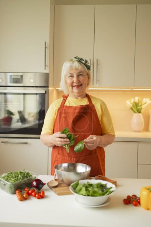 Senior 60s blond Caucasian smiling woman holding a knife and fresh spinach whole preparing a bowl of healthy diet salad in her rustic eco kitchen. High quality photo