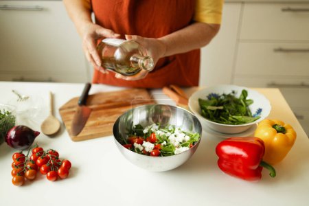 Frau würzt einen Gemüsesalat, während sie in der Küche Essen zubereitet. Der unkenntliche Koch fügt Olivenöl hinzu, während er zu Hause eine vegetarische Mahlzeit zubereitet. Konzept der gesunden Ernährung. 