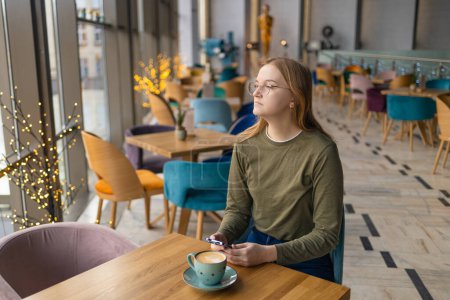 Mujer joven triste y pensativa que bebe café y mira su teléfono inteligente cerca de la ventana de la cafetería. Foto de alta calidad