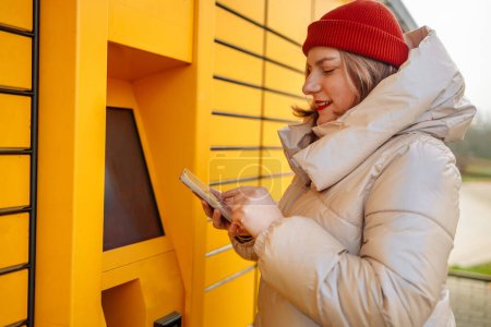 Femme recevant un colis de la boîte aux lettres automatique à l'aide d'un smartphone à l'extérieur. Concept de technologies de livraison modernes. Photo de haute qualité