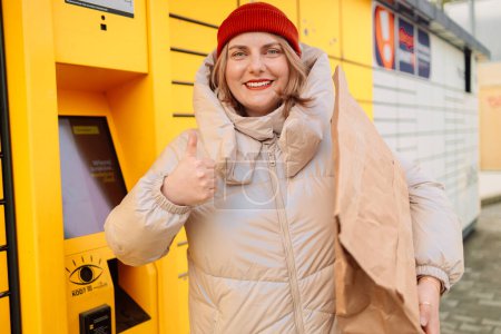 Frau empfängt Paket aus automatischem Briefkasten per Smartphone im Freien. Konzept moderner Liefertechnologien. Hochwertiges Foto