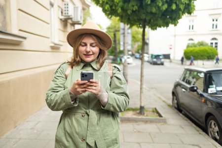 Portrait of Caucasian woman standing on street, city square and holding mobile phone. Girl using smartphone browsing internet. High quality photo
