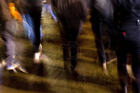 Téléchargez les photos : Photo avec caméra fait flou de mouvement d'une foule de jeunes gens marchant dans la ville pluvieuse la nuit - en image libre de droit