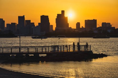 Téléchargez les photos : Vue depuis Odaiba, Tokyo, sur la baie jusqu'à l'horizon de Tokyo au coucher du soleil avec des silhouettes de personnes méconnaissables au premier plan - en image libre de droit