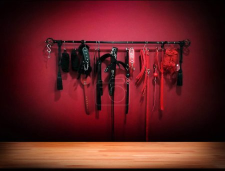 Foto de Mortage wood table top with whips for BDSM on red background on wall to present  Accessory for sexual games. - Imagen libre de derechos