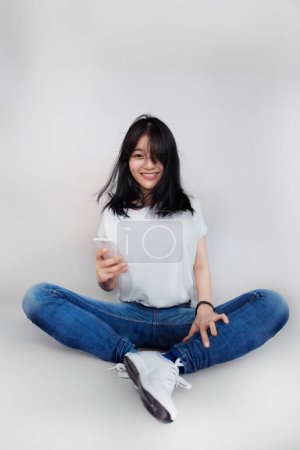 glücklich asiatische Mädchen mit Halter Werkzeug in lässig sitzen auf isoliertem Hintergrund und verwenden mobile auf isoliertem Hintergrund