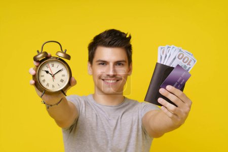 Photo for Young man smiles holding clock and wallet full of money on yellow background. Time is money concept and is ideal for use in financial planning, business, and success-related topics. Focus on hands - Royalty Free Image