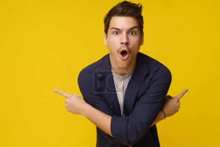 Photo for Surprised young man is depicted pointing with two crossed hands in opposite directions against yellow background, with plenty of copy space for text. Sense of astonishment, amazement, or shock, with - Royalty Free Image