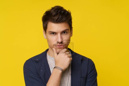 Photo for Young man on yellow background, holding his hand on his chin while deeply contemplating and thinking. Shot about intelligence, curiosity, and decision-making, as well as confusion, uncertainty, and - Royalty Free Image
