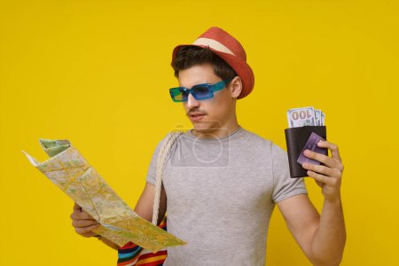 Photo for Young student is ready for journey, he looks into his travel map, holding his wallet with money and credit card. Yellow background with copy space perfect for travel-related designs and young - Royalty Free Image