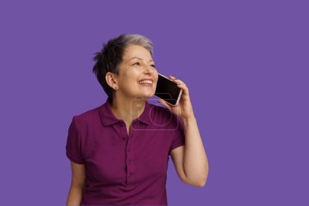 Téléchargez les photos : Femme âgée avec bonheur engage une conversation animée au téléphone. Isolé sur fond violet, son sourire rayonne de chaleur et de positivité, capturant l'essence de la communication joyeuse à l'ère numérique - en image libre de droit