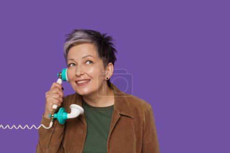 Photo for Senior lady engages in communication, holding old-fashioned wire phone to ear. Vintage charm, as senior lady connects with others against vibrant purple background. High quality photo - Royalty Free Image