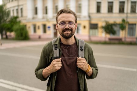 Photo for Man wearing glasses and carrying backpack, standing across city. Urban background sets scene for urban adventure, and mans appearance suggests he traveler ready to explore city. . High quality photo - Royalty Free Image