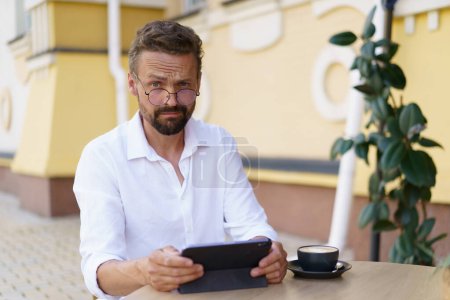 Photo for Funny man sits at table, engrossed in reading news on tablet PC. Face displays surprised expression, adding touch of humor to scene. Combination of the old town setting and modern technology. . High - Royalty Free Image
