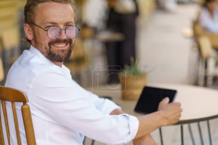 Téléchargez les photos : Homme d'âge mûr dans un moment franc assis à table dans un café. L'homme respire le sens de la beauté et de la confiance. Il peut être vu absorbé dans un PC de poche, un appareil portable qu'il tient dans ses mains. Haut - en image libre de droit