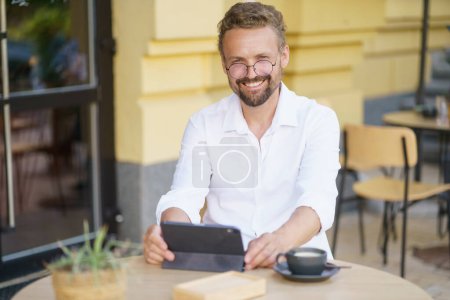Photo for White-collar worker enjoying some time outdoors at cafe. He seated at table, dressed in crisp white shirt. With cup of coffee by side, engrossed in reading and accessing information on tablet PC. - Royalty Free Image
