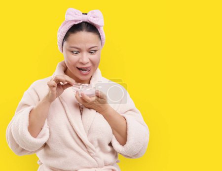 Téléchargez les photos : Mignon asiatique ménagère utiliser crème cosmétique sur fond jaune. Avec le sourire, elle se livre à une routine beauté, en appliquant de la crème sur la peau. Arôme de crème est si alléchant qu'il semble presque comme elle veut manger - en image libre de droit