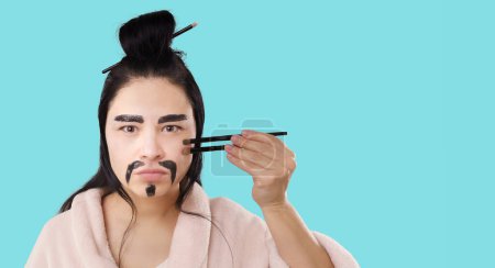Photo for Skin care and cosmetics with elements of Chinese kung fu and martial arts. Asian woman, face with painted mustache, hold brushes in chopsticks style to protect and defend her skin from age changes - Royalty Free Image