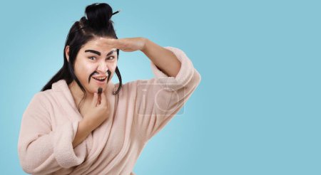 Téléchargez les photos : Femme asiatique avec moustache imite ludique combattant de kung fu chinois sur fond bleu. Femme au foyer Avec sens de l'humour peint les cheveux à la maison par elle-même. Photo de haute qualité - en image libre de droit