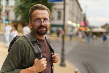 Téléchargez les photos : Homme d'âge moyen dans des lunettes dans des rues charmantes de la vieille ville. Avec un sourire amical et un comportement confiant, il tient le sac à dos, suggérant qu'il est voyageur ou aventurier explorant le paysage urbain historique. Haut - en image libre de droit