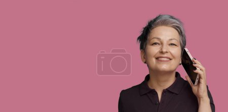 Photo for Successful Mature Woman Engages In Conversation On Mobile Phone, Isolated Against Pink Background. Concept Of Communication In Old Age, Showcasing Positive And Confident Use Of Technology By Seniors - Royalty Free Image