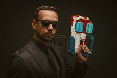 Téléchargez les photos : Un homme en costume pointu tient une arme à jouet, semblant imiter les actions d'un espion ou d'un agent de police. - en image libre de droit