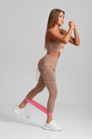 Téléchargez les photos : Fitness femme faisant de l'exercice pour les fessiers avec bande de résistance sur fond gris. Fille athlétique travailler - en image libre de droit