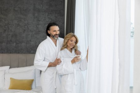 Foto de Hermosa pareja en albornoz en el dormitorio de pie cerca de las cortinas de la ventana - Imagen libre de derechos