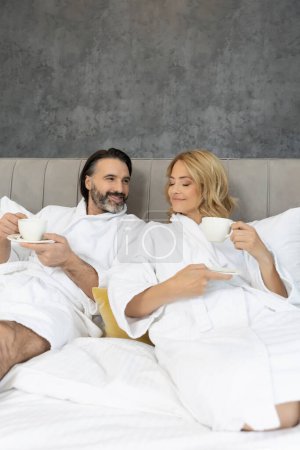 Foto de Hermosa pareja en albornoz en el dormitorio bebiendo café en la cama - Imagen libre de derechos