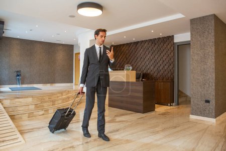 Foto de El hombre de negocios está teniendo una conversación en su teléfono inteligente mientras saca su maleta en el vestíbulo del hotel - Imagen libre de derechos
