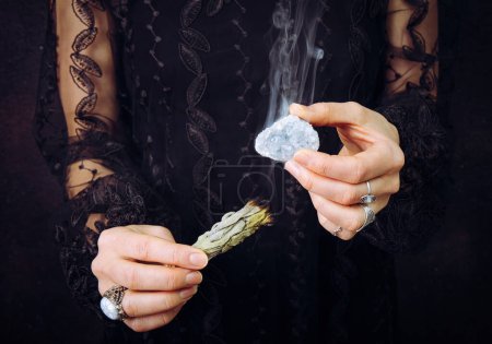 Vista de cerca de la mujer en vestido de encaje negro, limpieza de piedras preciosas de racimo de cristal celestita azul manchando paquete de salvia blanca. Eliminar la energía negativa, limpiar el cristal.