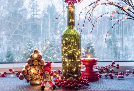 Weihnachtsdeko-Set mit Weinflasche gefüllt mit Mikro-LED-Partyleuchten und Fichte aus gebrauchten Weinkorken, niedliche Vintage-Elfenfigur, dahinter Fenster mit verschneitem Wald. 