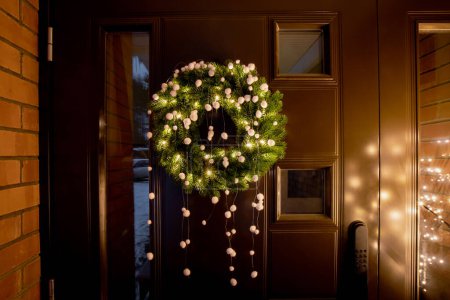 Grüne Farbe Fichtenspitzen Weihnachtskranz mit warmen Lichtern und weißen Filz Bommeln auf Hauseingang braune Metalltür mit winzigen Fenstern im Freien. Außerhalb der Weihnachtsdekoration.