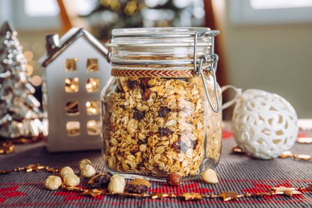Photo for Concept of homemade Christmas present, handmade roasted granola with various nuts and raisins in beautiful glass jar. Blur Christmas tree and lights on background. - Royalty Free Image