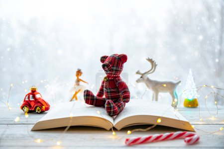 Open paper book with cute teddy bear shape figurine, fairy lights illuminated on white table, snowy forest landscape on background. Winter or Christmas children book reading concept.