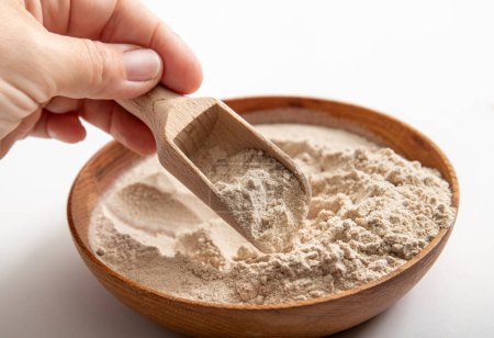 Person hand holding wood spoon and picking Psyllium husk flour powder from wood bowl indoors at home. Health benefits of Psyllium flour concept.