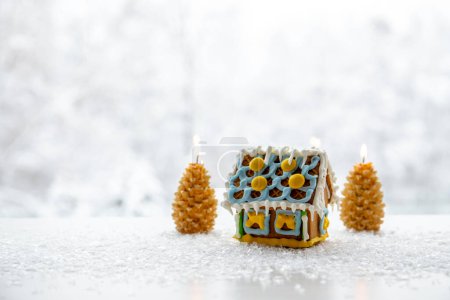 Téléchargez les photos : Bougies en forme d'épinette brûlant sur fond blanc enneigé avec paysage hivernal avec jolie maison en pain d'épice. Beaucoup d'espace pour copier. Hiver et Noël produits fond. - en image libre de droit