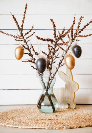 Photo for Tree branches in vase with black and bronze colored Easter eggs hanging on string, white rustic wood board background. Minimal home Easter decoration arrangement. - Royalty Free Image